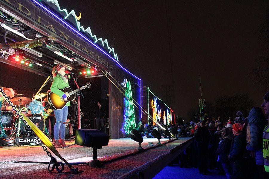 The Holiday Train opens up to a stage where performers sing holiday songs for the audience. The bright lights and decorations on the Holiday Train create a festive atmosphere.