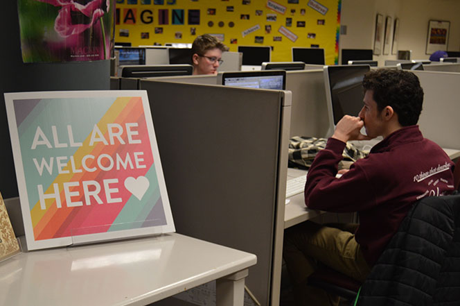 Senior Simon Lewin works on a computer  in the Library Media Center next to the All Are Welcome Here sign. Media Specialist Ellen George put the sign to send a message of acceptance and understanding. 