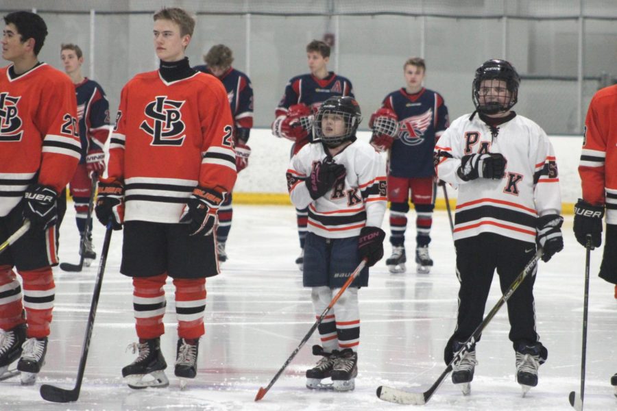 Junior Jacob Johnson and other team members stand along with the game kids during the Pledge of Allegiance.  Each game the team picks one or two kids who play for St. Louis Park and allow them to skate around on the ice before the game with the team. 
